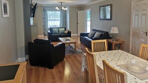 Uma sala de estar com sofá, mesa de centro, mesa de jantar com cadeiras, luminária e uma janela grande com cortinas.