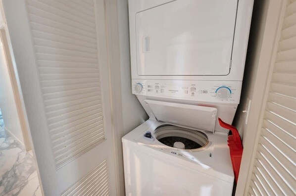 Een kleine wasruimte met een voorlader wasmachine en droger, een bedieningspaneel met diverse instellingen, en een rode slang.
