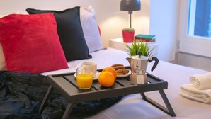 A neatly made bed with red and black pillows, a bedside table with a lamp, a small plant, and a tray with orange juice, cookies, and a coffee pot. - Casa King - Welchome (Cannobio)