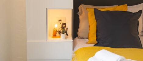 A neatly made bed with a yellow and white blanket, a rolled white towel, and a black pillow.