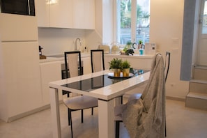 Une cuisine moderne avec des meubles blancs, une table à manger au plateau en verre et une vue sur l’extérieur par une fenêtre.