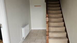 Una escalera con peldaños alfombrados en color beige y barandillas de madera, que conduce a un rellano con un radiador blanco y un interruptor de luz en la pared.