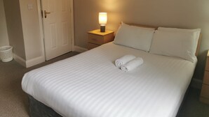 Una cama bien hecha con sábanas blancas, una lámpara de noche, una mesita de madera y una puerta a la izquierda.