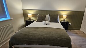 Una cama bien hecha con una manta verde, dos lámparas de noche y una ventana con persianas.