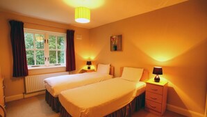 A hotel room with two single beds, a window with curtains, a radiator, a nightstand with a lamp, and a small drawer. - Bunratty West No 40026 (Bunratty)
