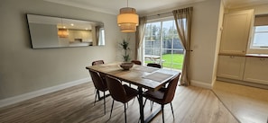 A dining area with a wooden table, six chairs, a hanging light fixture, and a large window with curtains.