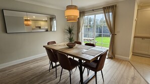 A dining area with a wooden table, six chairs, a hanging light fixture, and a large window with curtains.