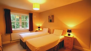 A hotel room with two single beds, a window with curtains, a radiator, a nightstand with a lamp, and a small drawer. - Bunratty West No 40023 (Bunratty)