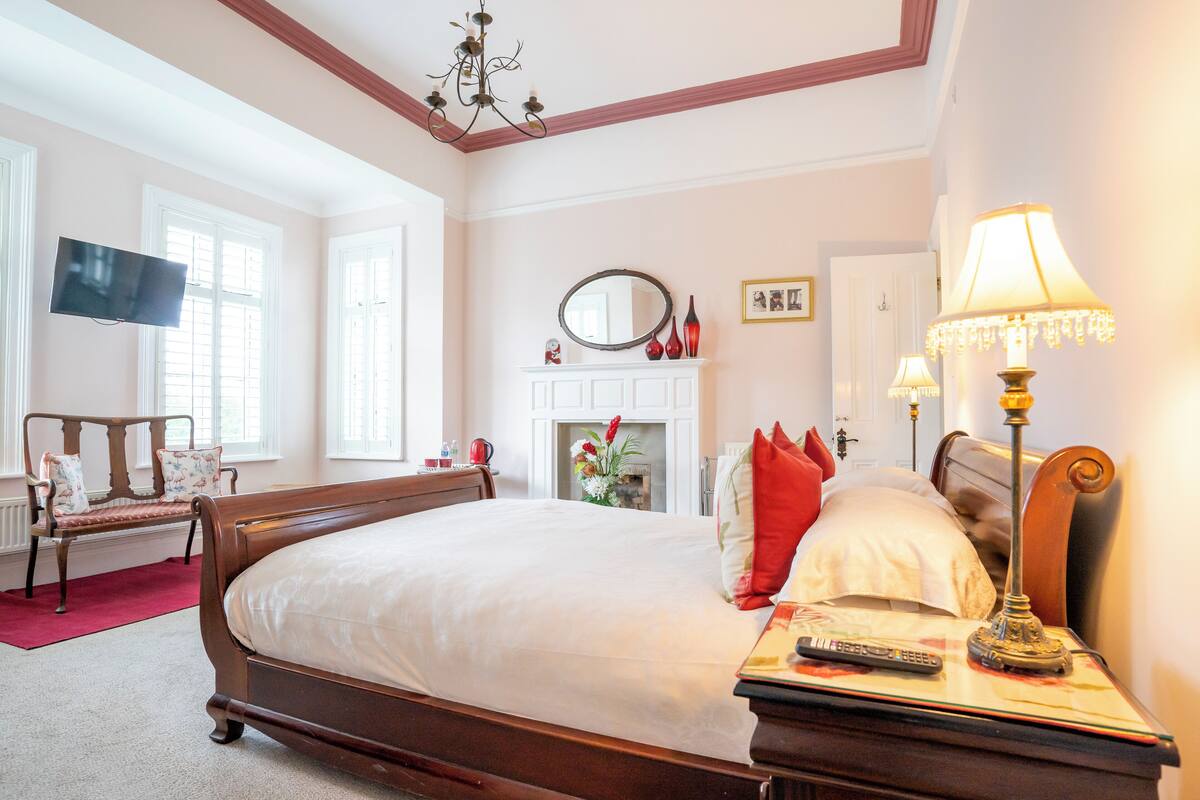A spacious bedroom with a large bed, a television mounted on the wall, a fireplace, and a nightstand with a lamp.
