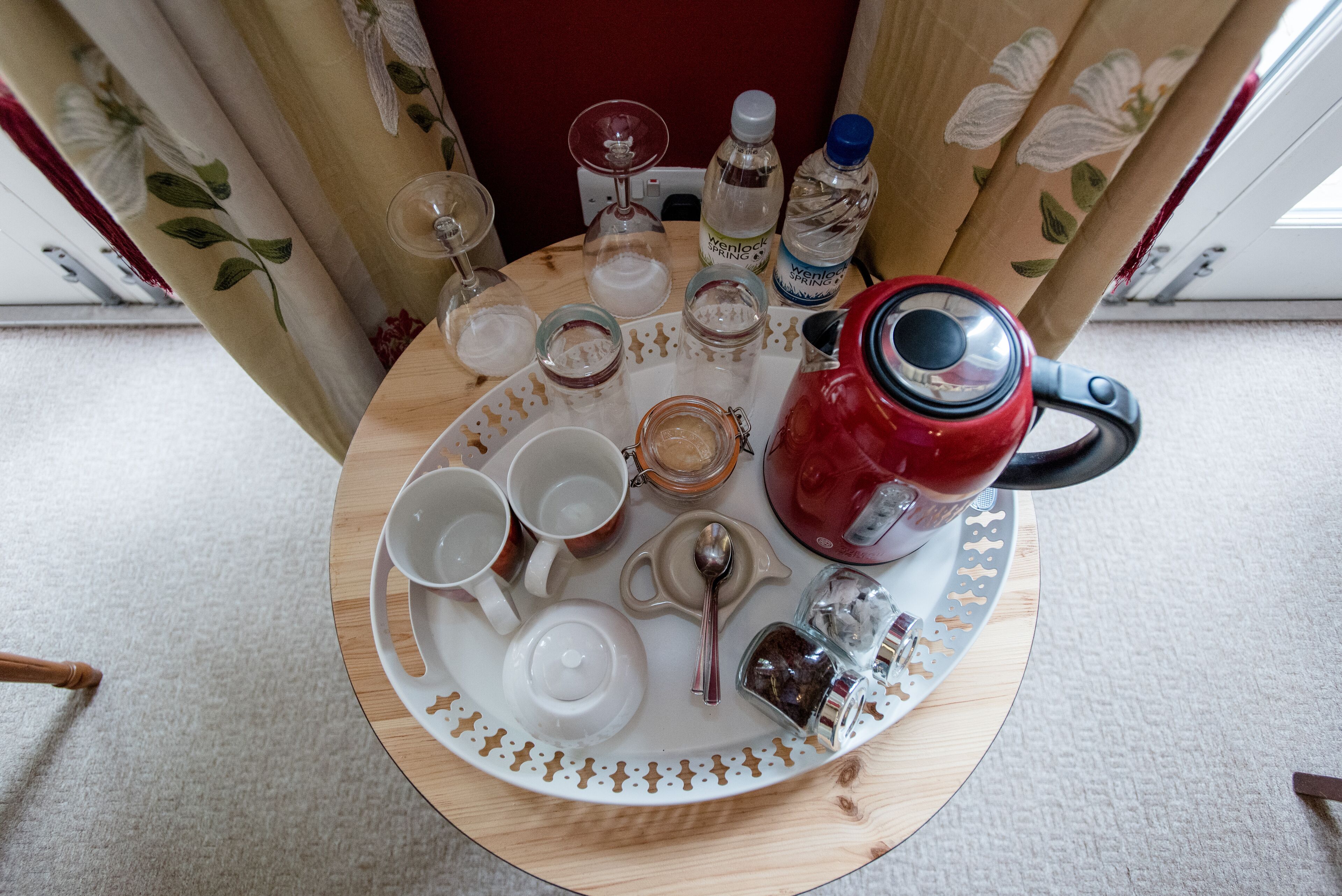A round wooden table with a white tray holding various items including a red kettle, cups, a spoon, and bottles.
