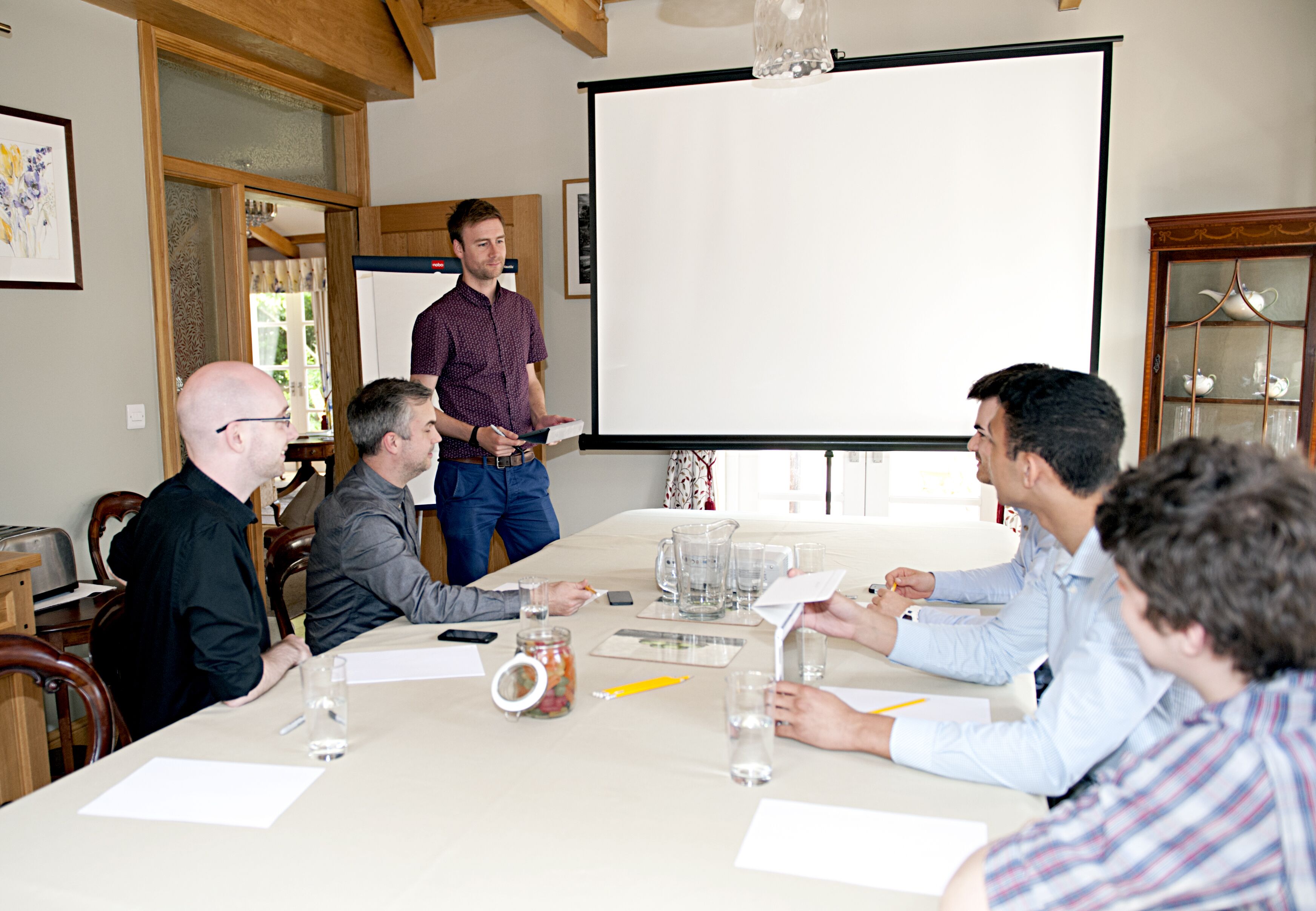 A meeting room with a large table, chairs, a projector screen, and various items like a jar, glasses, and papers.