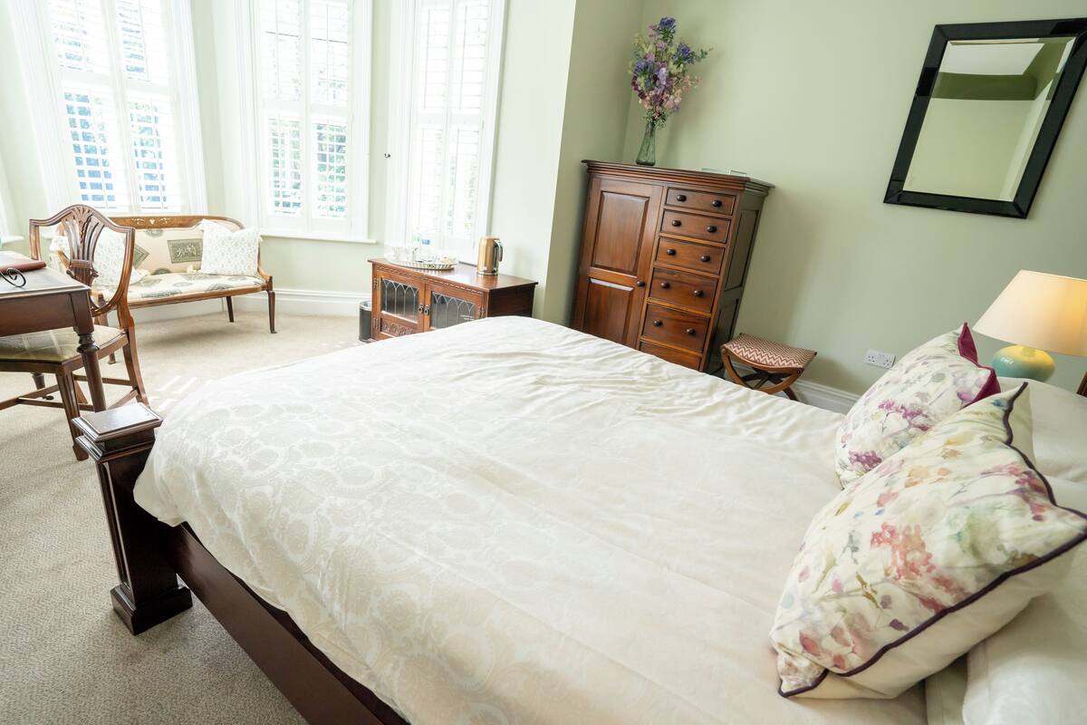 A neatly arranged bedroom with a large bed, a wooden dresser, a chair, and a desk.