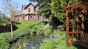 A garden with a pond, a wooden bench, and a brick house in the background. - Mere Brook House (Wirral)