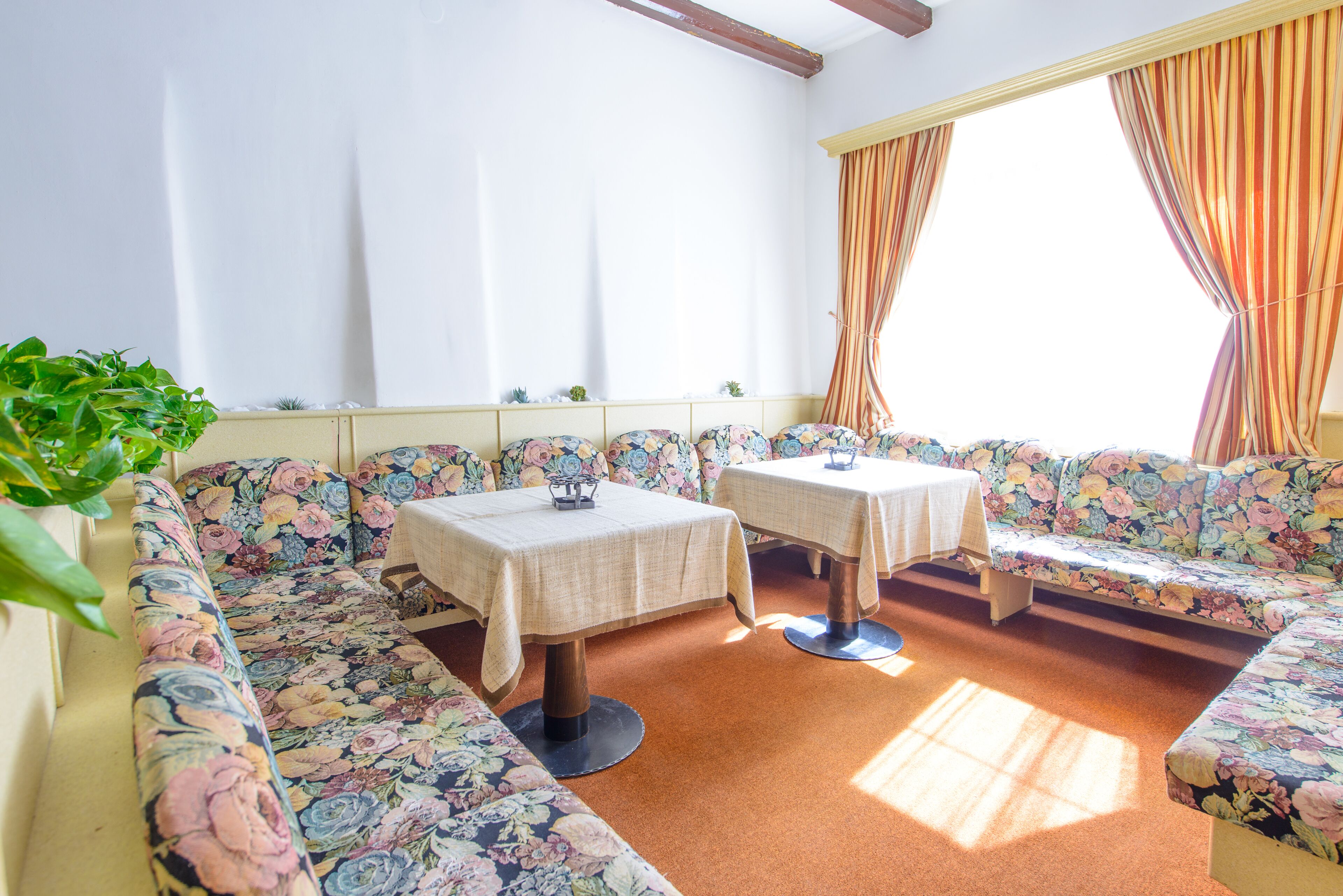 Ein Zimmer mit einer blumigen Couch, zwei kleinen Tischen mit weißen Tischdecken und einem Fenster mit Vorhängen.