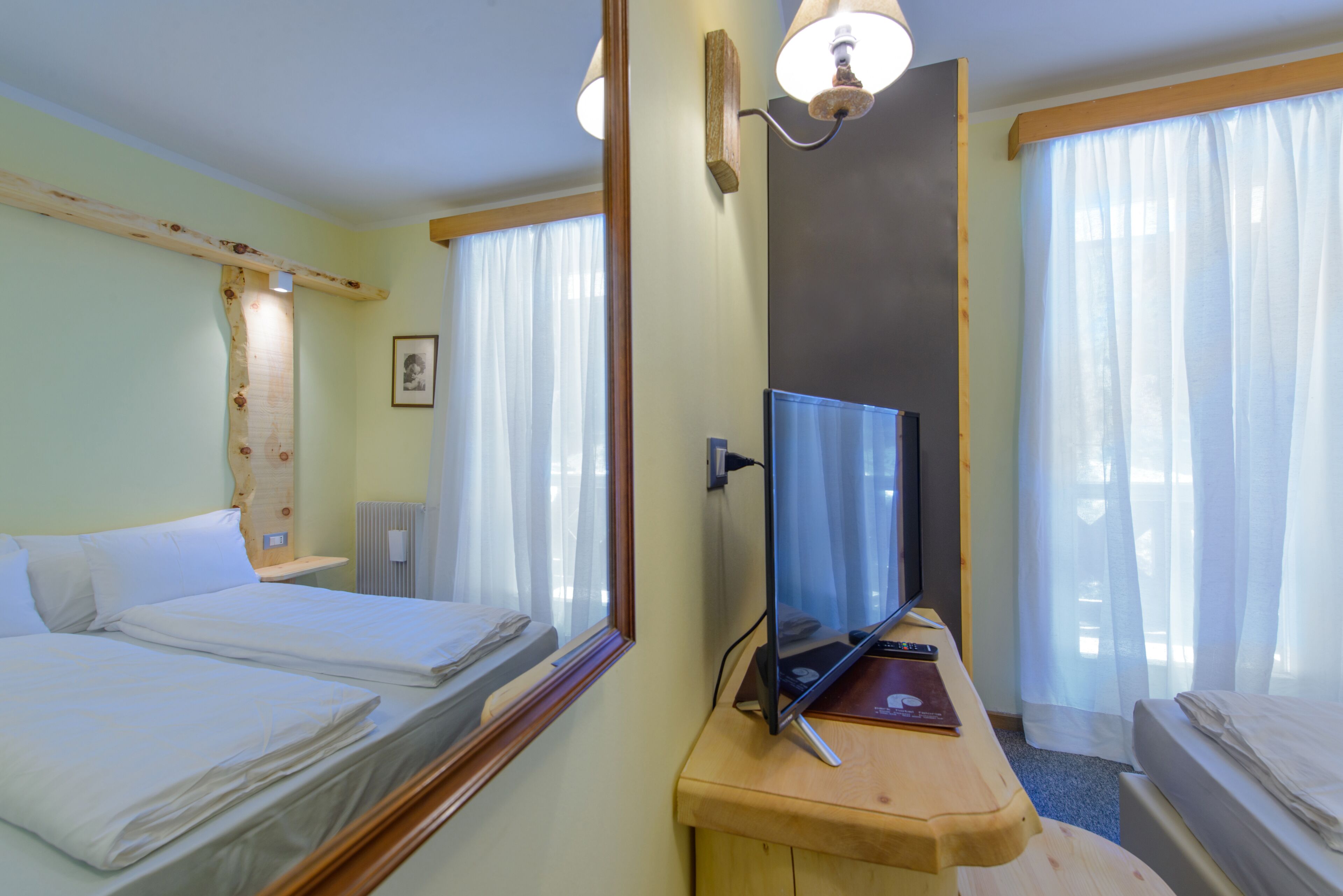 A hotel room with two beds, a TV, a wooden desk, and a large window with sheer curtains.