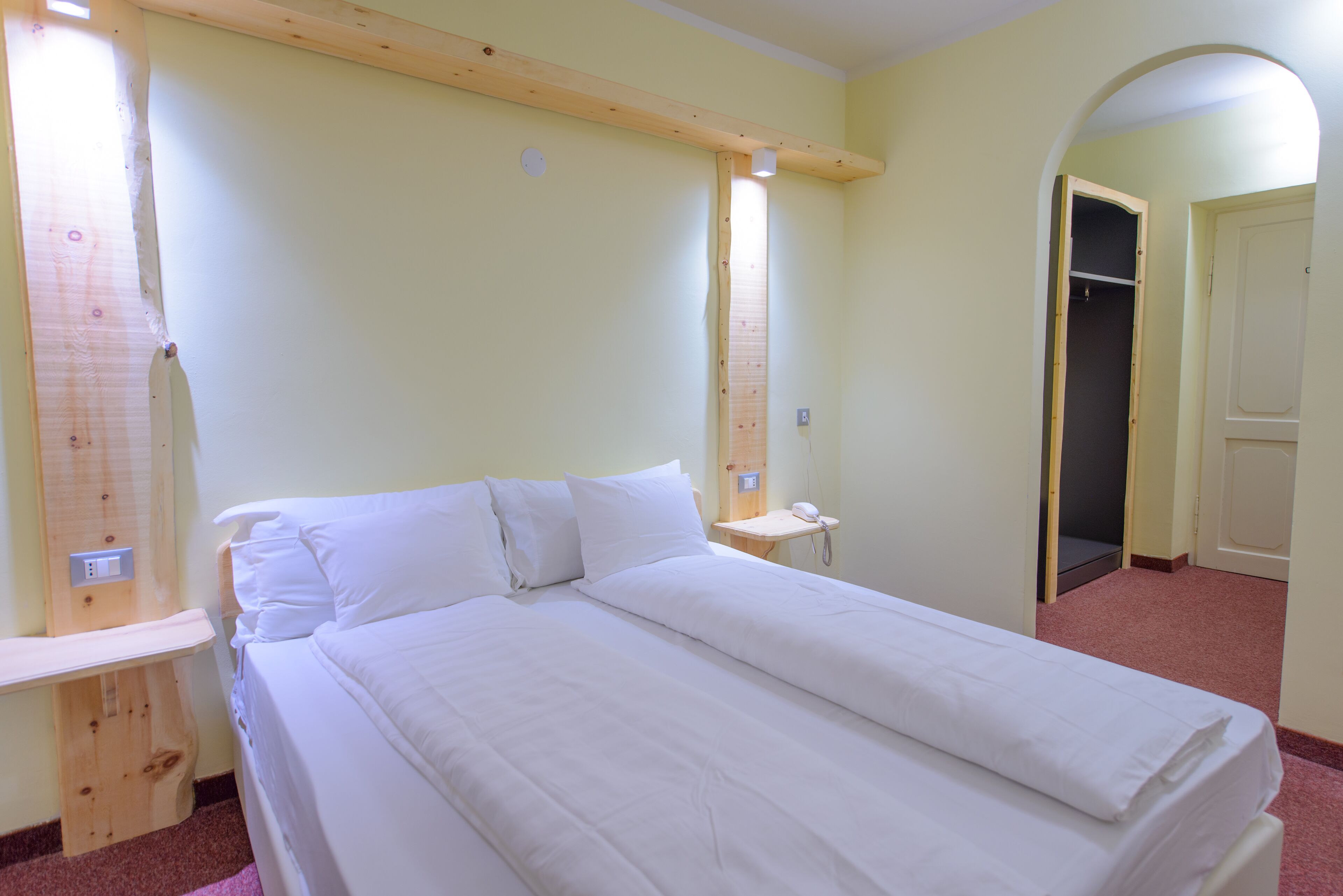 Una habitación con una cama grande, una mesita de noche de madera, una pequeña mesa con un teléfono y una puerta que da a otra habitación.
