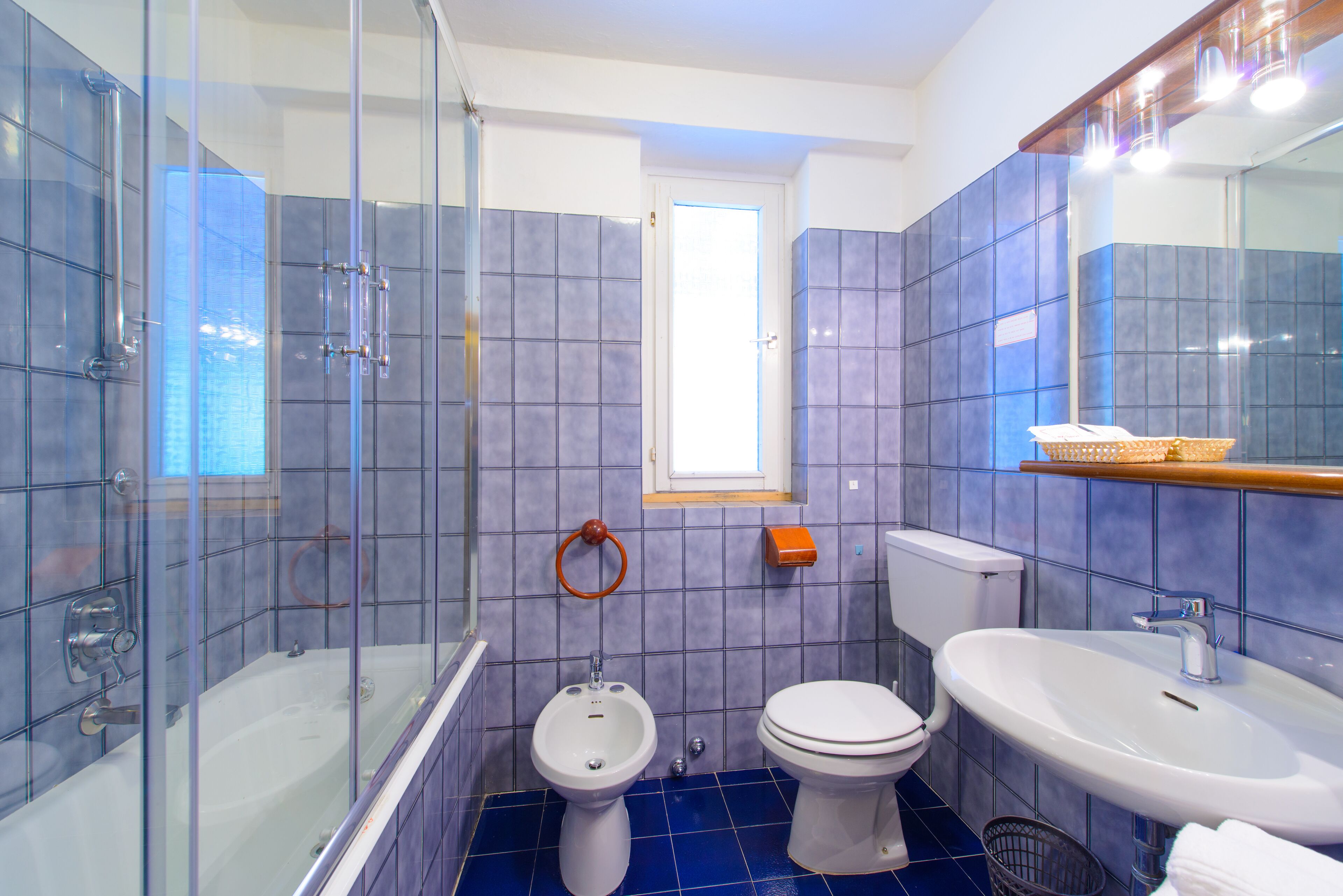 A bathroom with a glass-enclosed shower, two toilets, a sink, and a window.