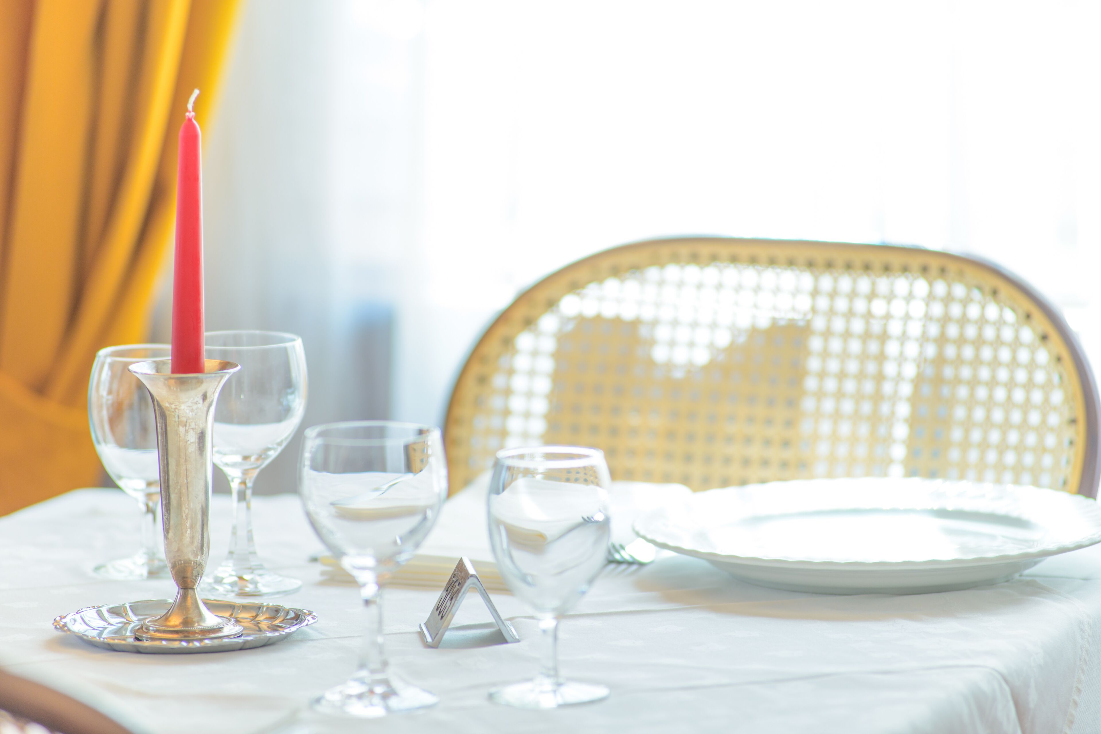 Una mesa de comedor dispuesta con cristalería transparente, una vela roja y un plato blanco, con una silla de mimbre al fondo.