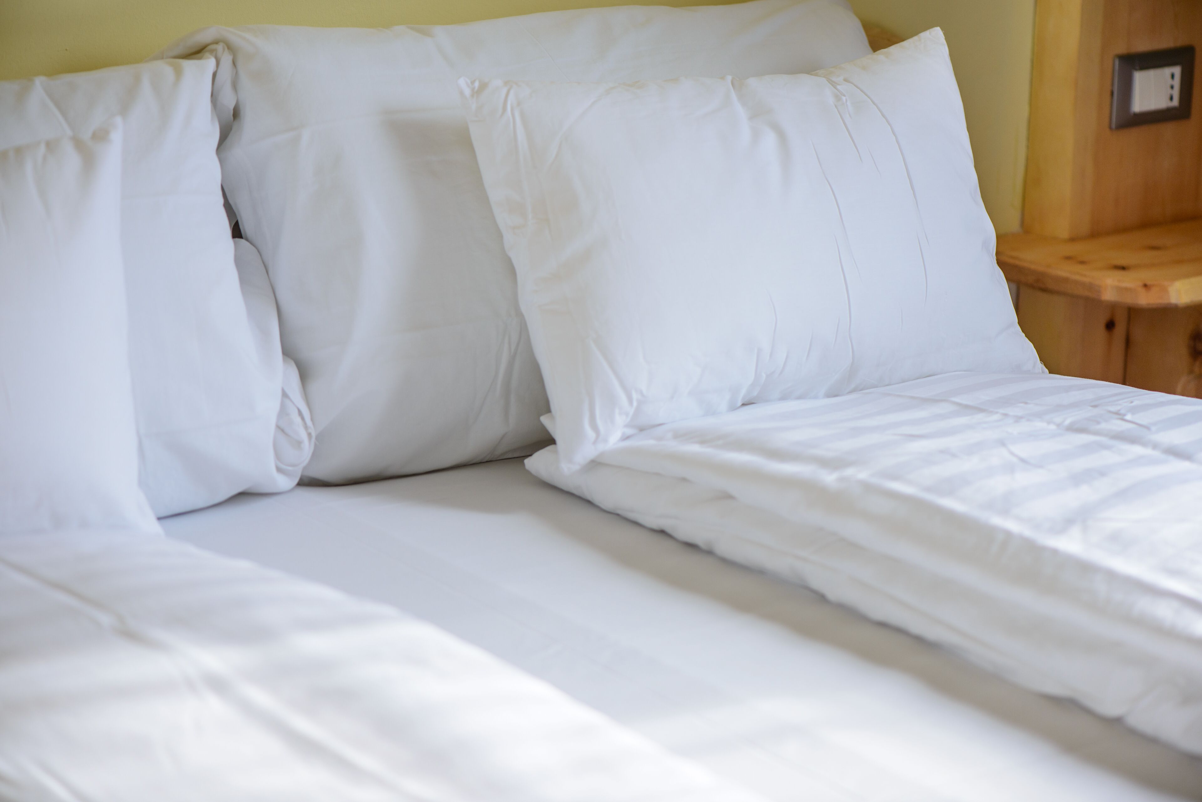A bed with white linens and pillows, a wooden nightstand, and a wall-mounted electrical outlet.