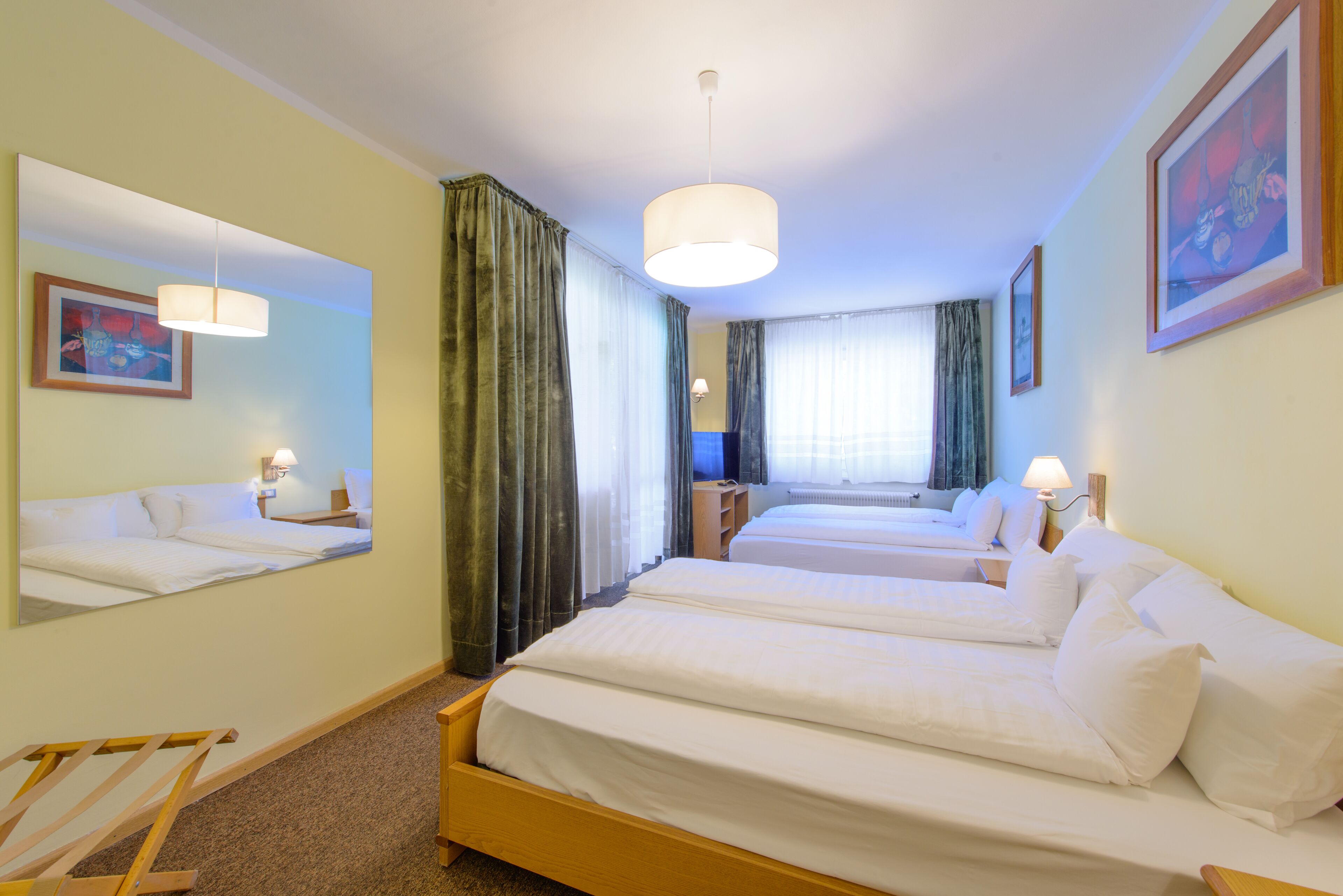 A hotel room with two single beds, a large mirror, a wooden chair, and a window with curtains.