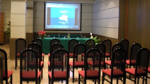 Una sala conferenze con uno schermo di proiezione che mostra il nome di un hotel, un tavolo con un microfono e file di sedie.