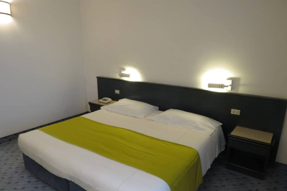 A hotel room with a bed, two bedside tables, a nightstand, and a wall-mounted light fixture.