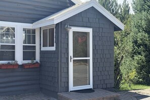 En liten, envåningsbyggnad med en grå fasad, en vit dörr och en liten veranda med krukväxter.