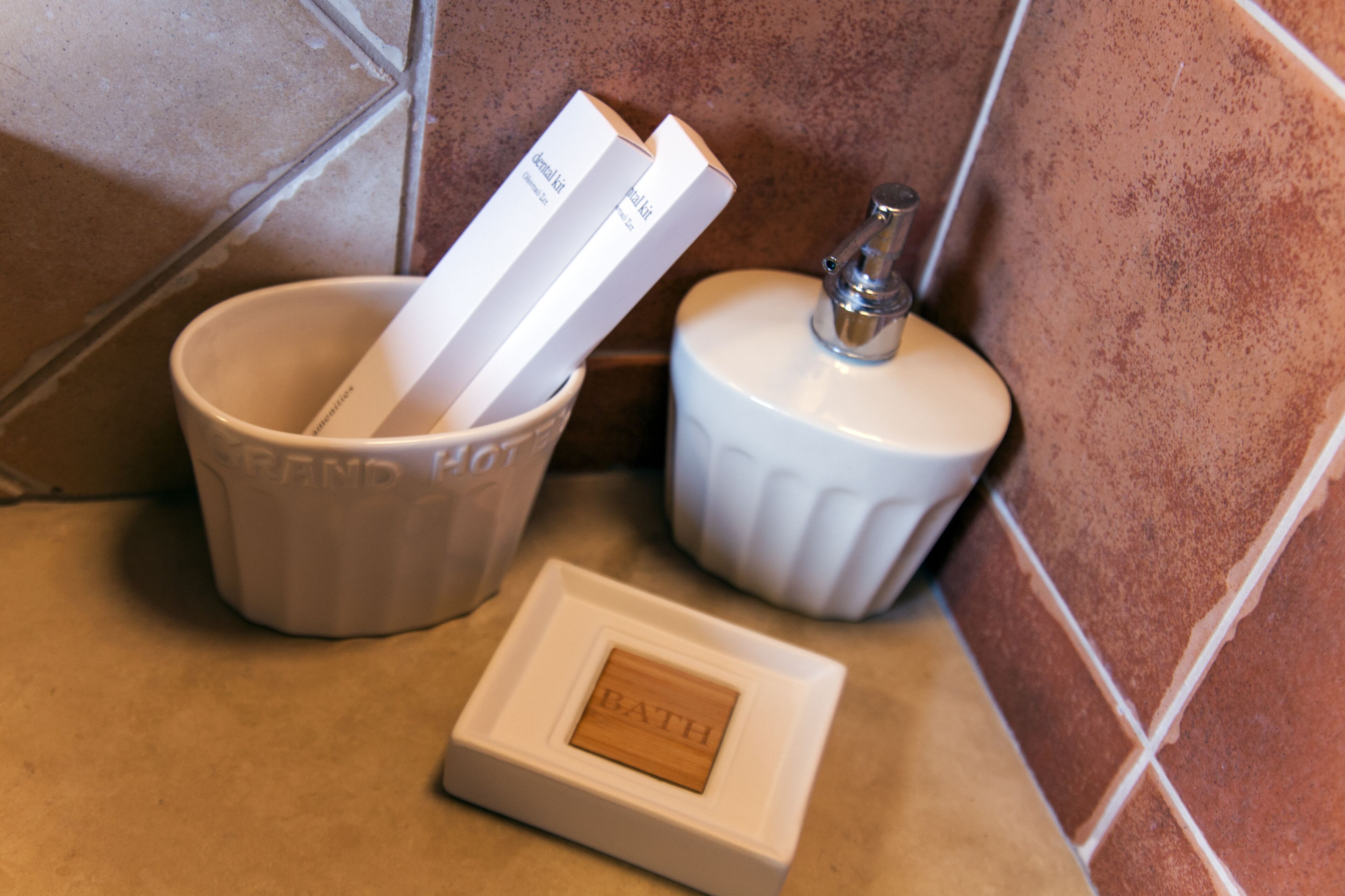 Un coin de salle de bain avec un lavabo blanc contenant deux flacons blancs, un porte-savon blanc et une petite boîte en bois sur laquelle est inscrit le mot « BAIN ».