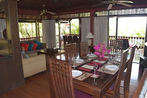 A dining area with a long table set for six, surrounded by wooden chairs. There is a sofa with colorful cushions in the background, and a decorative vase with pink flowers on the table.