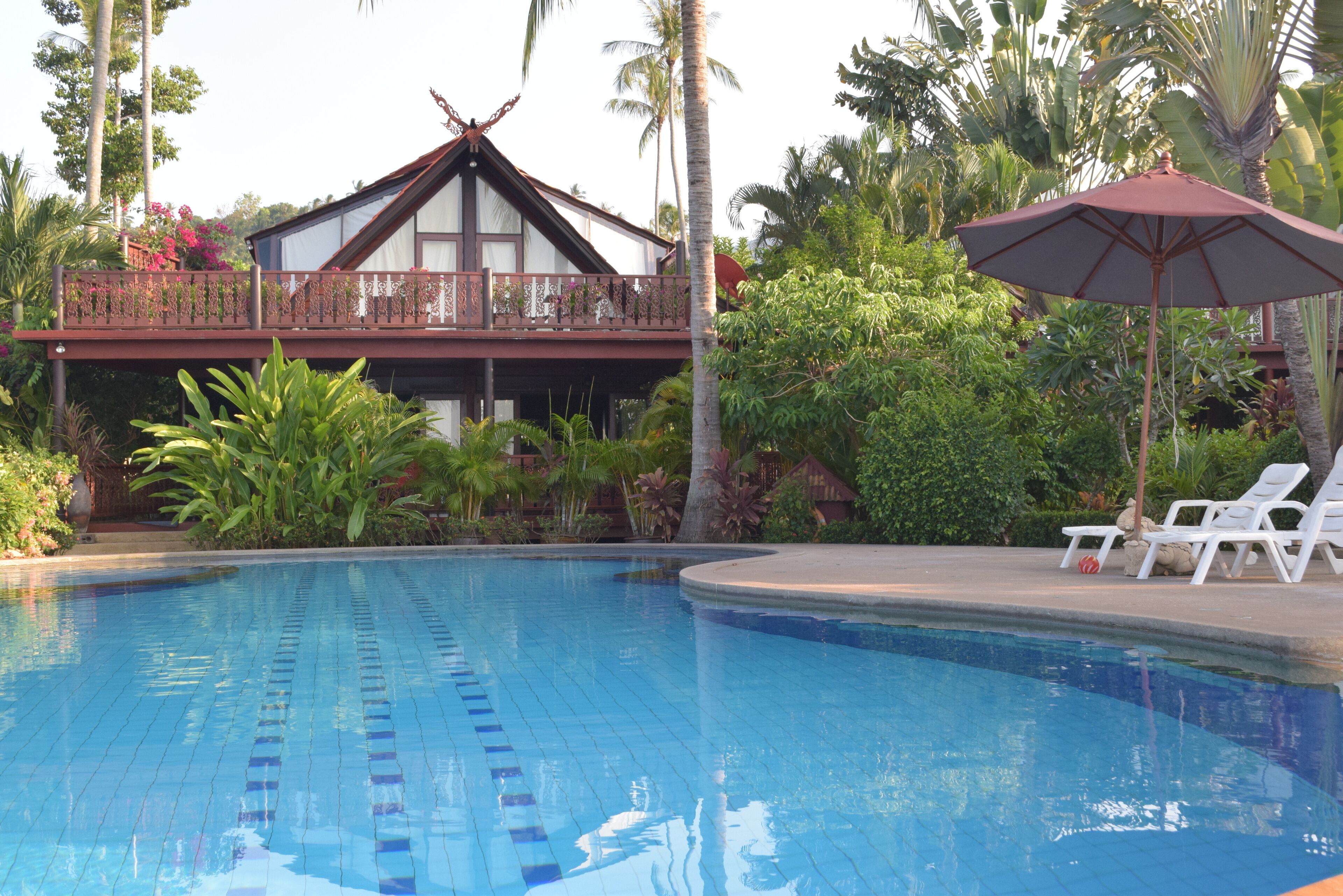 A swimming pool with clear blue tiles, a wooden deck, and a lounge area with white chairs and a red umbrella.