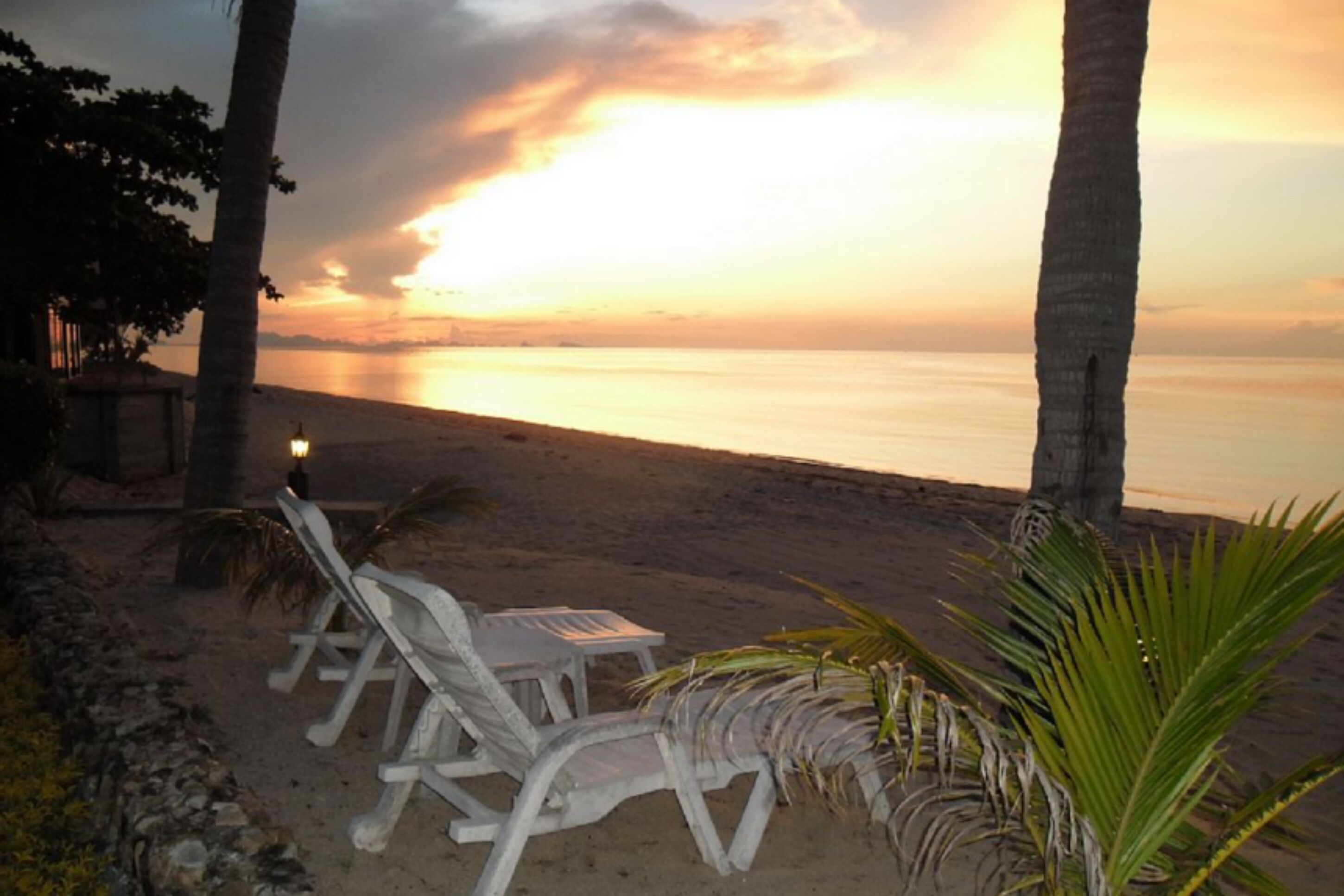 Una playa con sillones blancos, una mesa pequeña, palmeras y una puesta de sol sobre el agua.
