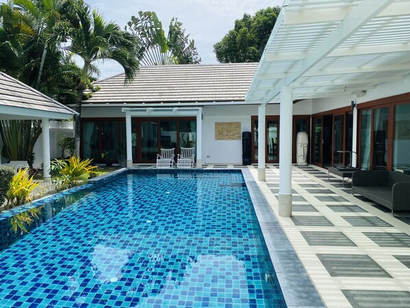 A swimming pool with a tiled floor, surrounded by a covered patio area with chairs and a statue.