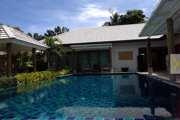 A swimming pool with a tiled deck, surrounded by a covered patio area with chairs and a table.