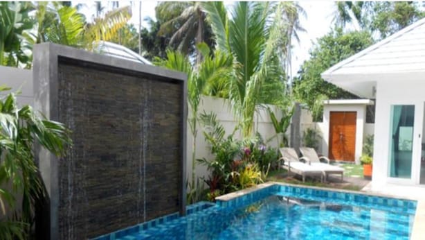 En swimmingpool med kakelbeklädd kant, omgiven av frodig grönska och ett modernt hus.