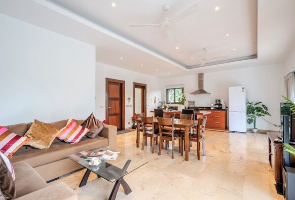 Amplia sala de estar con sofá, mesa de centro, mesa de comedor y sillas. Se observa una cocina con refrigerador y estufa al fondo.