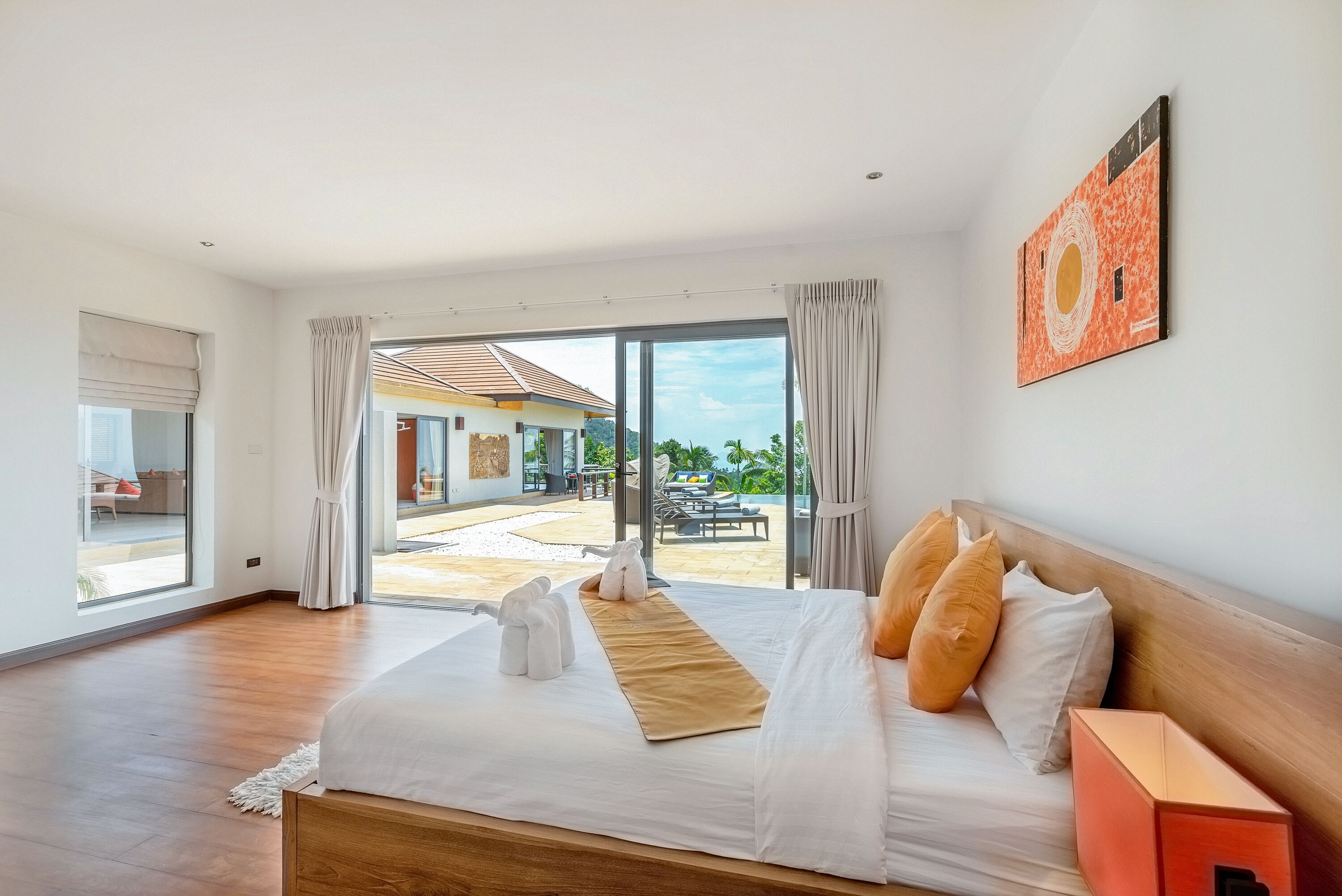 A spacious bedroom with a large bed, a view of a pool and outdoor seating area, and a modern art piece on the wall.