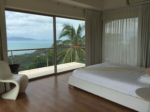 Une chambre avec une grande fenêtre, un lit, une chaise et une vue sur la mer et les palmiers.