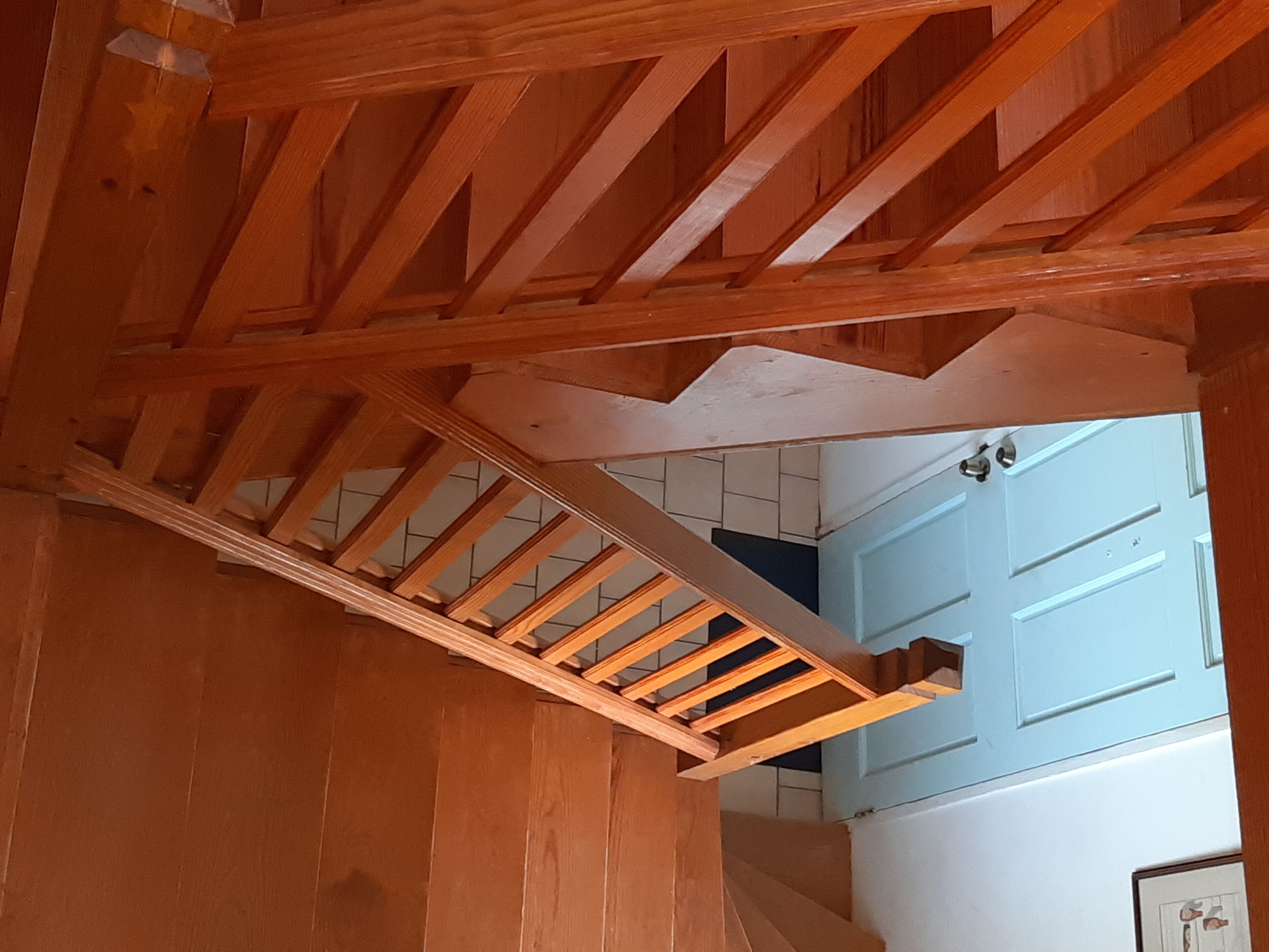 Un escalier en bois avec une rampe, menant à une pièce dont la porte est bleue et les murs blancs.