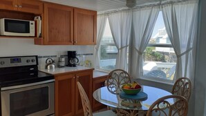 Una cocina compacta con armarios de madera, microondas, estufa y cafetera. Hay una mesa de vidrio redonda con sillas y un bol de fruta.