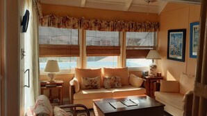 Una sala de estar acogedora con un sofá, dos sillones, una mesita de centro y una lámpara. Hay tres ventanas con persianas, un ventilador de techo y cuadros enmarcados en la pared.