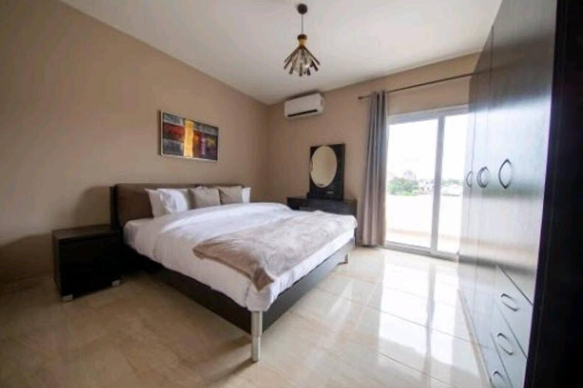 A modern bedroom with a large bed, a nightstand, a ceiling light, and a view of the outdoors through a large window.