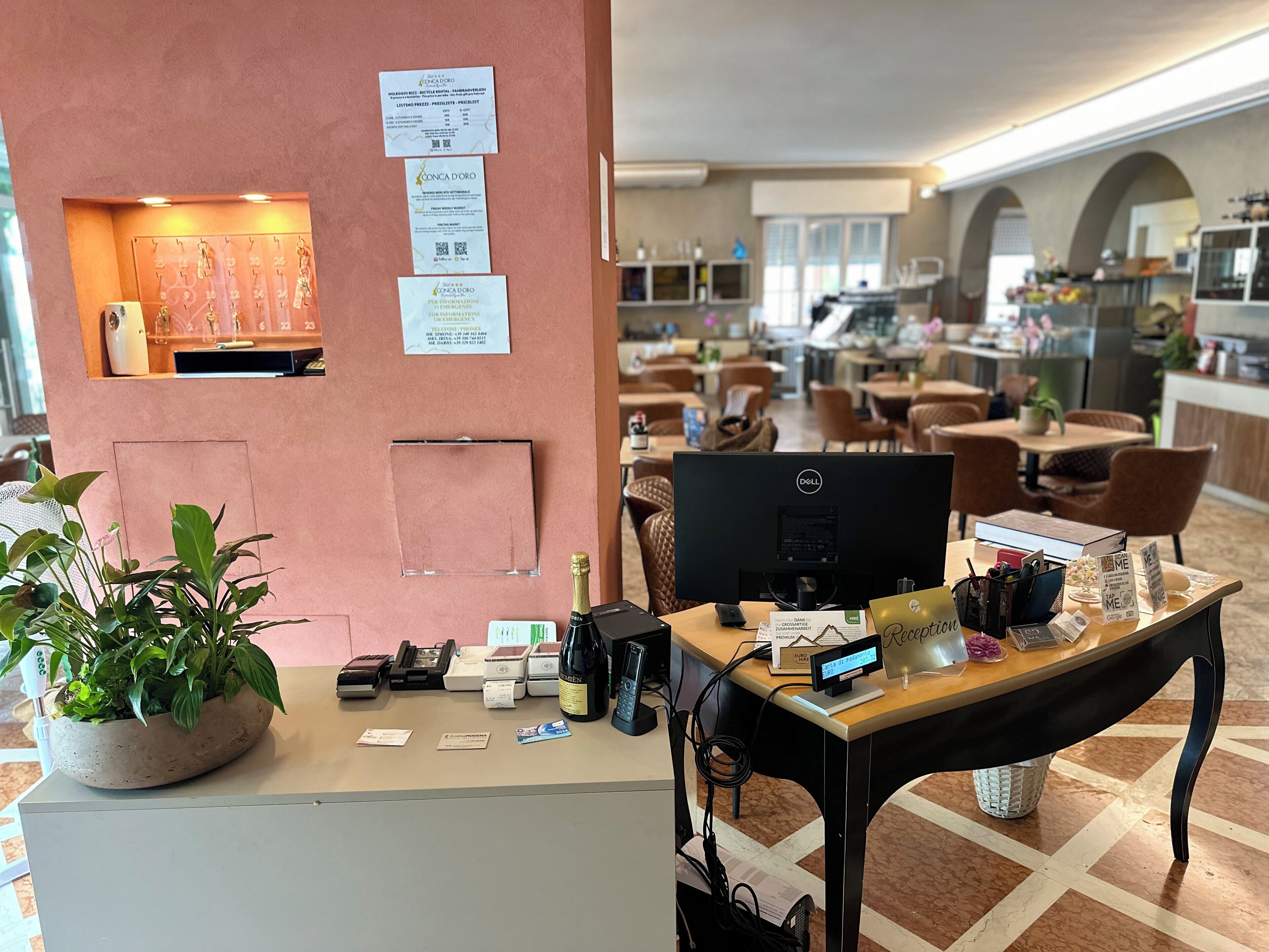 Ein Hotelrezeptionstresen mit Computer, Telefon und verschiedenen Dokumenten. Links steht eine Pflanze und an der Wand ein Schild.