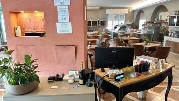 Ein Hotelrezeptionstresen mit Computer, Telefon und verschiedenen Dokumenten. Links steht eine Pflanze und an der Wand ein Schild.