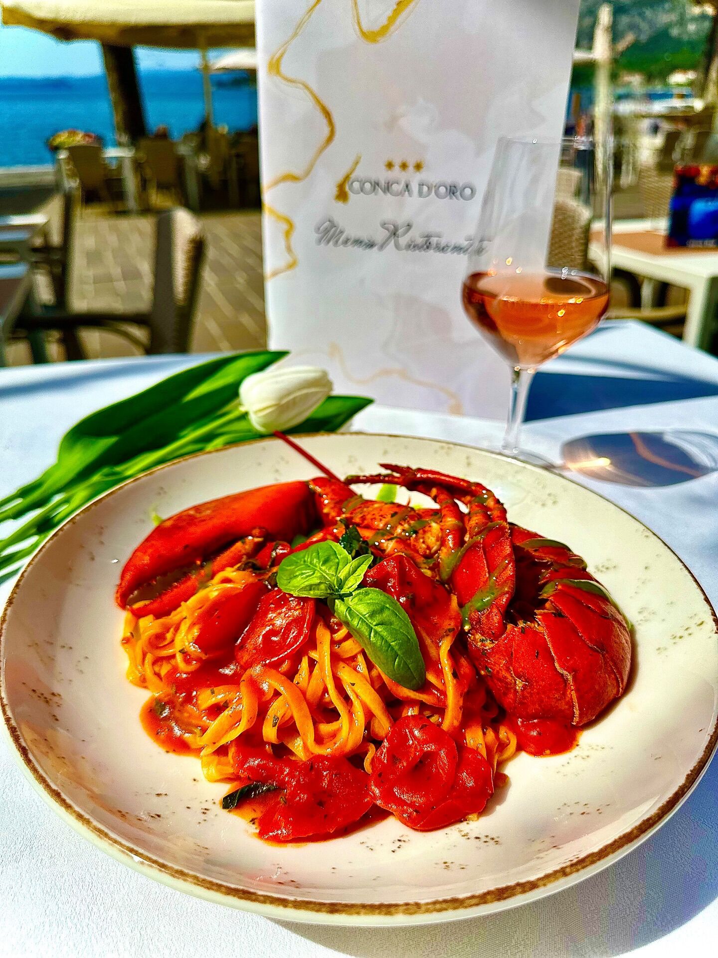 A seafood pasta dish with a glass of rosé wine and a menu card on a table by the sea.