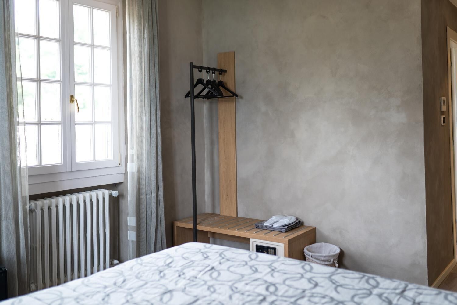 A bedroom with a bed, a radiator, a wooden bench with a phone, a coat rack, and a window with curtains.
