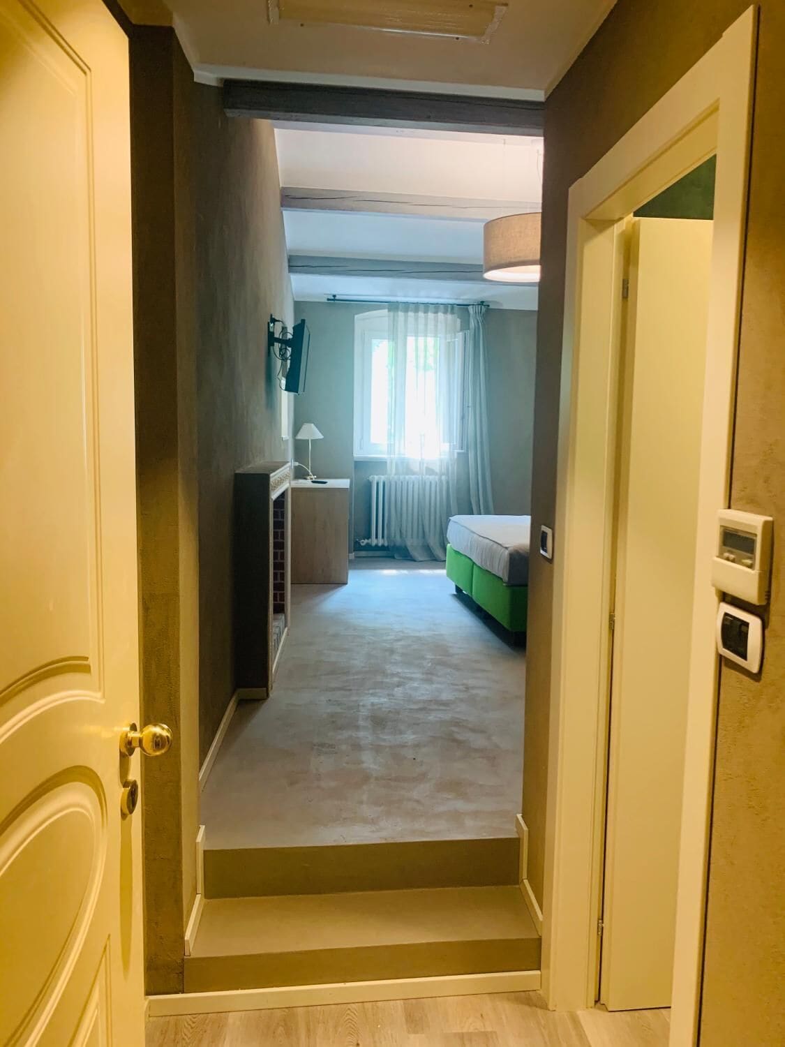 Un couloir menant à une chambre à coucher équipée d’un lit, d’un radiateur et d’une fenêtre avec des rideaux.