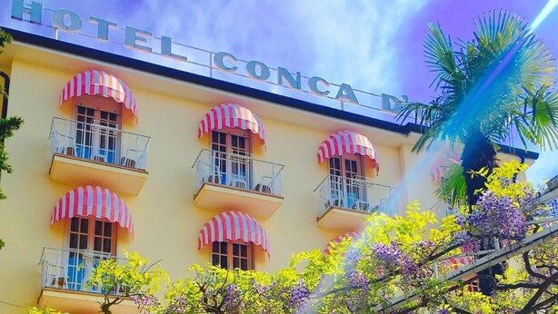 Un hôtel appelé « HOTEL CONCA D », doté de nombreux balcons, chacun étant surmonté d’un auvent à rayures.