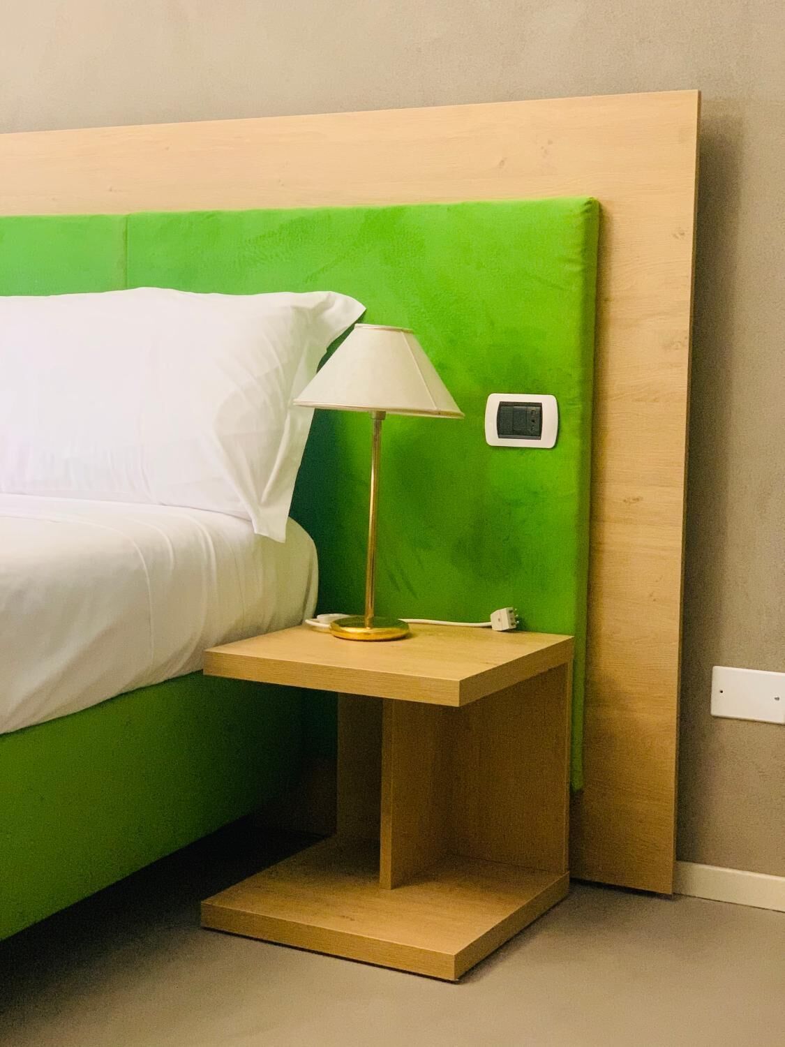 Una cama con cabecera verde, una mesita de noche con lámpara y un enchufe en la pared.