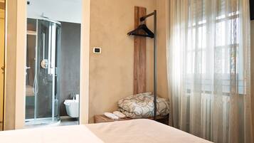 A hotel room with a bed, a shower, a toilet, and a window with sheer curtains.