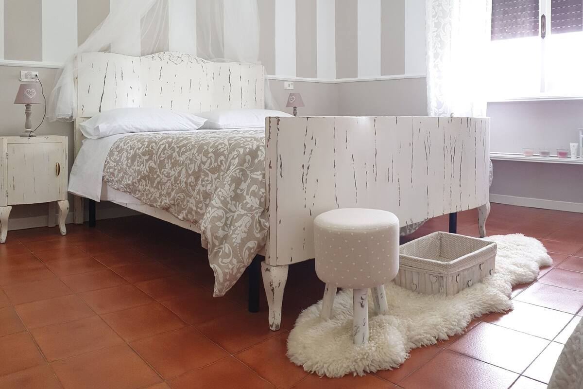A bedroom with a bed, a nightstand, a small stool, a white air conditioning unit, and a window with curtains.